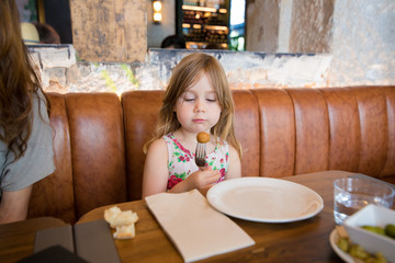Four years age blonde girl eating and looking at croquette in fork next to woman mother sitting in brown leather sofa at restaurant
