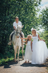 Man, woman dressed as a bride, girl and white horse in the park among the green trees