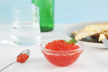 Red caviar and toast of white bread. Caviar in a glass cup, toast on a plate. Next a glass with water and a bottle, a spoon with caviar. Light background. Close-up. Macro photography.