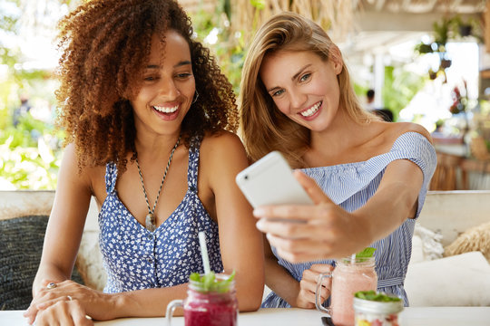 Mixed race female friends make pictures on cell phone, use special application. Two beautiful smiling women take selfie on cellular during free time on weekend at cozy restaurant. Recreation time