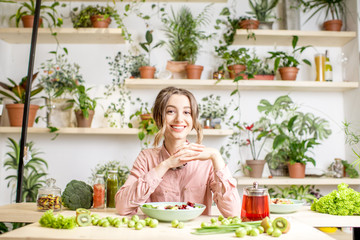 Portrait of a young woman sitting with healthy food in the beautiful interior with green flowers on the background