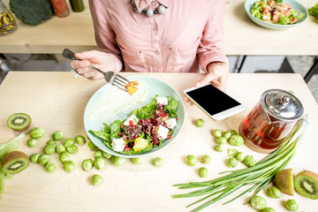 Woman eating salad and using smartphone at the table dwcorated with green food ingredients. Top view