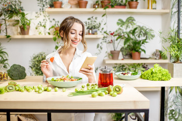 Obraz na płótnie Canvas Young woman eating healthy food sitting with smartphone in the beautiful interior with green flowers on the background