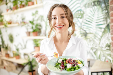 Young woman eating healthy food sitting in the beautiful interior with green flowers on the...