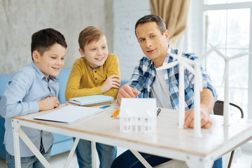 Clear explanation. Cheerful pleasant young father sitting at the table with his two sons and explaining them the construction of wind turbines