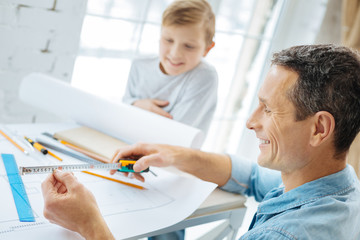 Obraz premium Important measures. Pleasant pre-ten boy sitting at the table in his fathers office and watching him check the measures on the tape while working on the blueprint