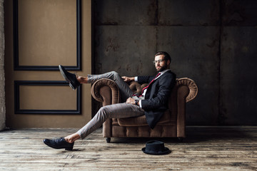 handsome elegant businessman posing in armchair, hat lying on floor near, loft interior