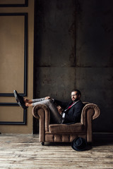 elegant businessman posing in armchair, hat lying on floor near, loft interior