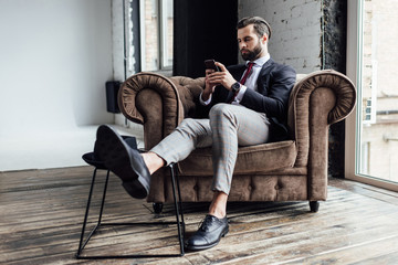 stylish businessman using smartphone and sitting in armchair in loft