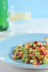 Tar-tar from salmon with avocado and pomegranate seeds on a blue plate. In the background a glass with water and a bottle. Light background. Close-up.  Vertical orientation of the frame.