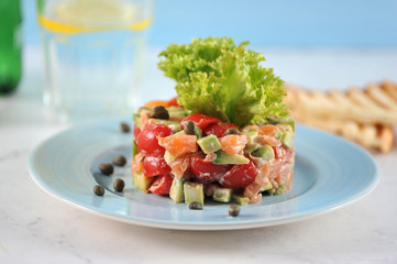 Tar-tar from salmon with avocado and tomatoes on a blue plate. In the background, toast, a glass of water. Light background. Close-up. Macro photography.