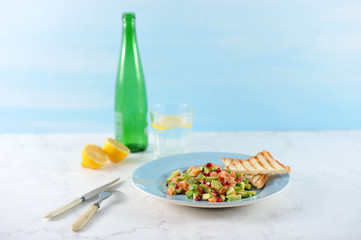 Tar-tar from salmon with avocado and pomegranate seeds on a blue plate. On a plate toast. There are cutlery nearby. In the background, a lemon, a glass of water and a bottle. Light background. 