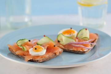 Sandwiches with salmon. The sandwich is decorated with fresh cucumber slices, hard-boiled egg, onion rings. In the background a glass with water. Light background. Close-up. 