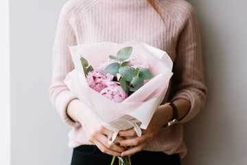 Young woman holding a small beautiful blossoming peonies flower bouquet on the grey wall background, cropped photo