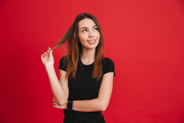 Portrait of young smiling woman 20s wearing black looking upward and touching brown strand with brooding or dreaming view isolated over red background