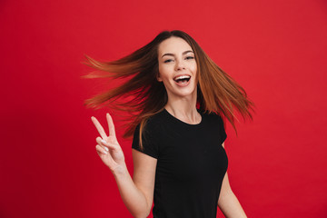 Image of positive woman with long brown hair posing on camera with showing peace sign, isolated over red background