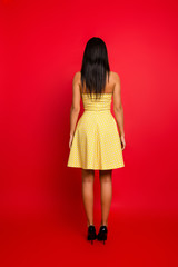 Vertical full-size rear view portrait of charming beautiful pretty woman wearing yellow dotted dress with naked shoulders, isolated on red background