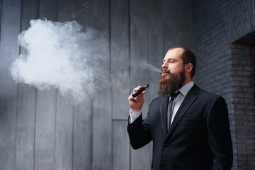 Elegant vaper. Handsome young bearded man in suit smoking vaporizer electronic cigarette.