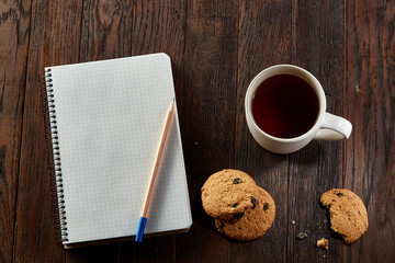 Cup of tea with cookies, workbook and a pencil on a wooden background, top view
