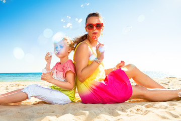 happy trendy mother and child on seashore blowing bubbles