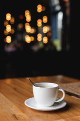 A white coffee cup with a spoon inside on a wooden table of a cafe, bokeh lights in the background.