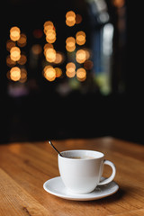 A white coffee cup with a spoon inside on a wooden table of a cafe, bokeh lights in the background.