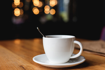 A white coffee cup with a spoon inside on a wooden table of a cafe, bokeh lights in the background.