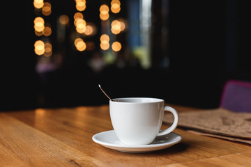 Obraz premium A white coffee cup with a spoon inside on a wooden table of a cafe, bokeh lights in the background.