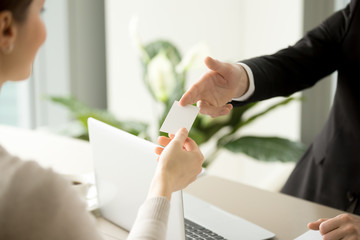 Company male manager giving blank business card over work desk with laptop to female client or business partner. Business leaders exchanging their contacts on meeting in office. Back view. Close up