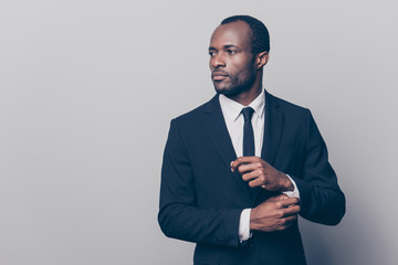 Portrait of stunning, trendy, attractive, dreamy, perfect man in black suit with tie fasten button on sleeve cuffs of white shirt, looking to the side, isolated on grey background - Powered by Adobe