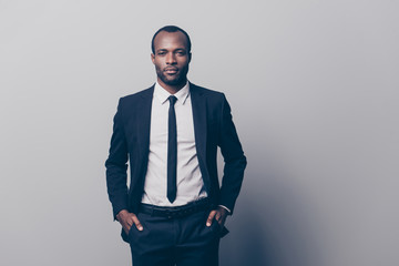 Portrait of stunning, manly, brutal man in black tux, tuxedo with white shirt and tie, holding two hands in pockets of pants, looking at camera, going for meeting, isolated on grey background