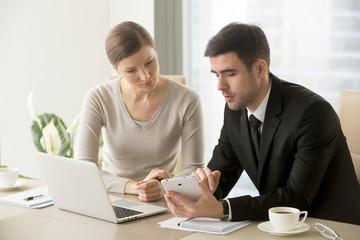 Millennial businessman showing project calculations, concept design, drawings, financial indicators to female colleague or business partner on tablet screen while sitting at desk with laptop in office
