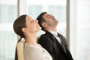 Satisfied relaxed businesswoman and businessman resting while sitting on chairs with closed eyes, taking short break for meditative practice during day in office. Office workers imagining success