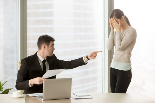 Serious male executive manager with bad financial report in hand blaming inexperienced female employee in mistakes at documents, stressed and upset young woman cowering her face with hands and crying