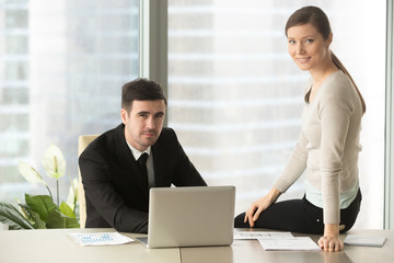 Fototapeta premium Candid portrait of millennial male and female business leaders sitting together at office desk with laptop and papers and looking at camera with smile. Positive company coworkers posing at workplace