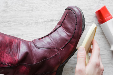 Woman's hand applying transparent shine cream on red leather boots with sponge. Cares about boots beauty and protection from cold and wet. Preparing for winter season. Footwear cleaning concept.