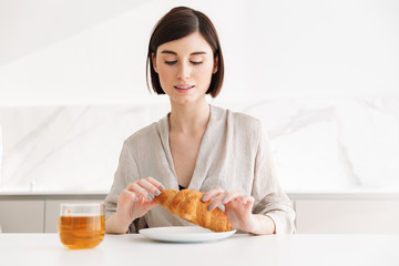 Photo of adorable morning woman having breakfast in hotel room, and enjoying croissant with tea