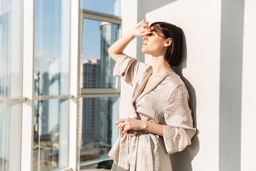 Young gentle woman with short dark hair in housecoat standing sunlit near window, and covering face in sunny morning