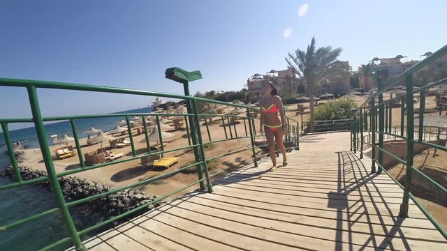 Pretty girl goes on a wooden bridge in the resort and cute smiles. In the background there is a blue sea and many umbrellas from the sun.