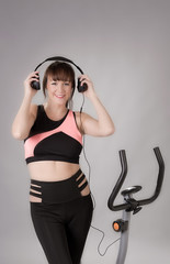 Fototapeta premium Woman holding headphones with an exercise bicycle against a grey background
