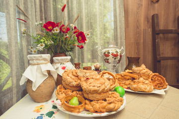 Traditional Russian baked goods. Table in rustic house, on which there are plates with buns, pies and pretzels, samovar, clay jugs with milk, and vase with bouquet of summer flowers