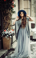 Smiling woman in hat and sweater stands in light dress on a winter street before the building decorated for winter holidays