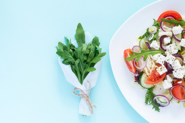 Fresh spring salad with cucumber, tomato, cheese and arugula isolated on a white plate Bouquet of fresh green arugula leaves in white  craft paper