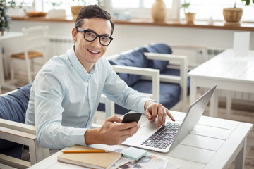 Obraz premium Freelancer. Good-looking glad dark-eyed dark-haired man smiling and working on his laptop and holding his phone while sitting at the table