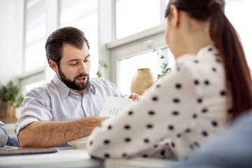 Obraz premium Showing results. Handsome concentrated bearded man holding a sheet of paper and showing a diagram to his partner sitting opposite him