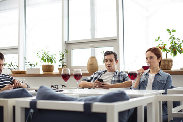 Fototapeta premium Weekend. Handsome serious dark-haired young man using his phone and his friends sitting at the table with him and drinking wine