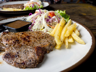 close up of T-bone steak with salt and pepper, golden french fries and green vegetables. on white plate, Fork and knife, wooden table in the restaurant