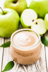 Baby food, applesauce in glass jar and green apples on shabby white wooden background. Selective focus, space for text. 