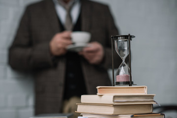 close-up shot of stack of books and hourglass with man drinking coffee blurred on background