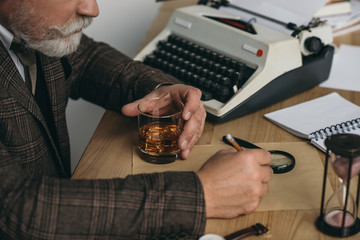 cropped shot of senior writer working on manuscript and holding glass of whiskey